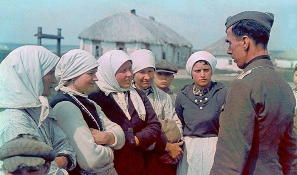 A soldier speaks with village women and children beside thatched cottages