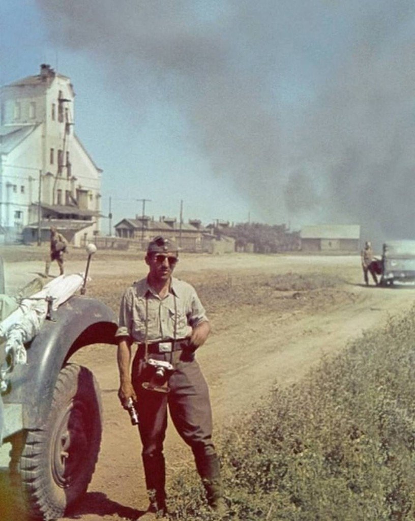 Soldier with a camera stands on a dusty road as smoke rises behind him